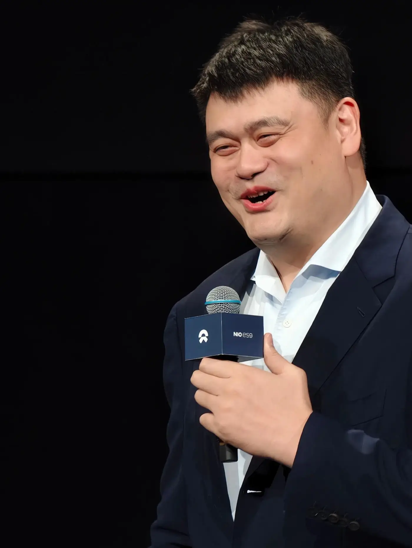 Yao Ming in a dark suit, holding a microphone and smiling broadly at a public event