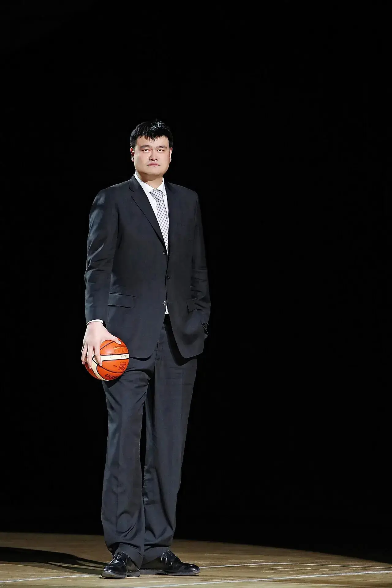 Yao Ming standing alone on a basketball court in a dark suit, holding a ball at his side